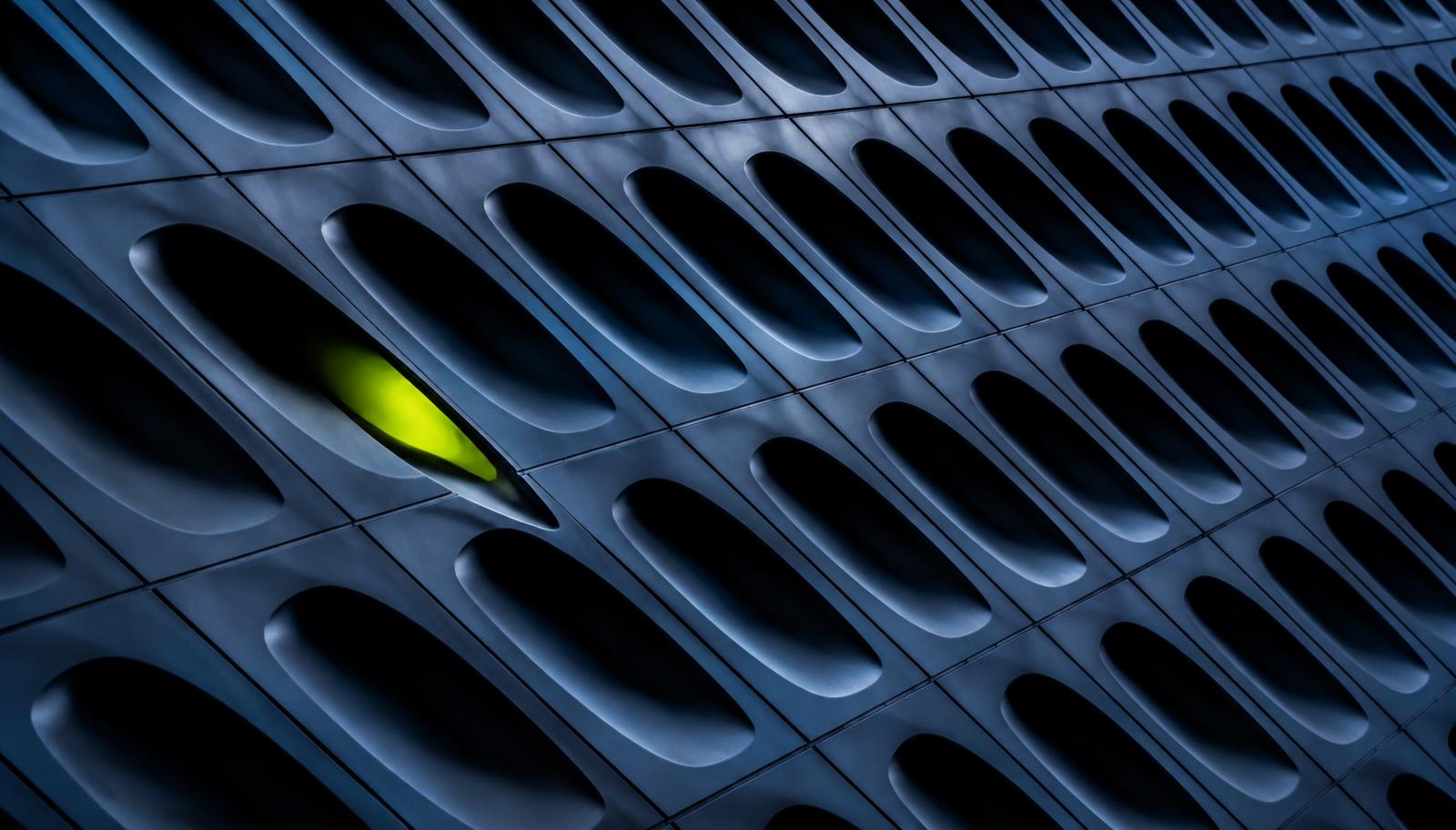 Abstract close-up of a dark blue engineered facade of repeating apertures with one lit in lime green
